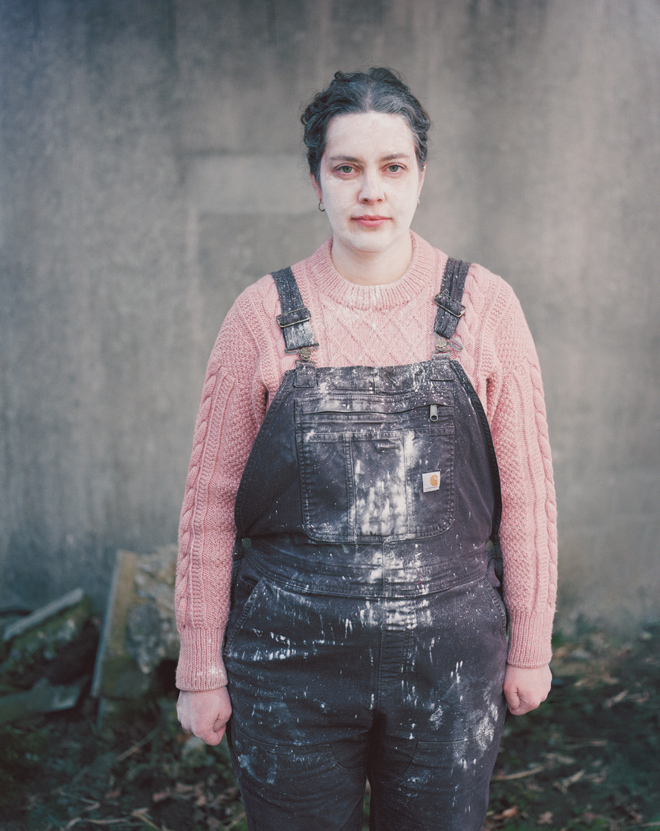Woman in pink sweater and black overalls stained with white dust standing outdoors against a gray wall.