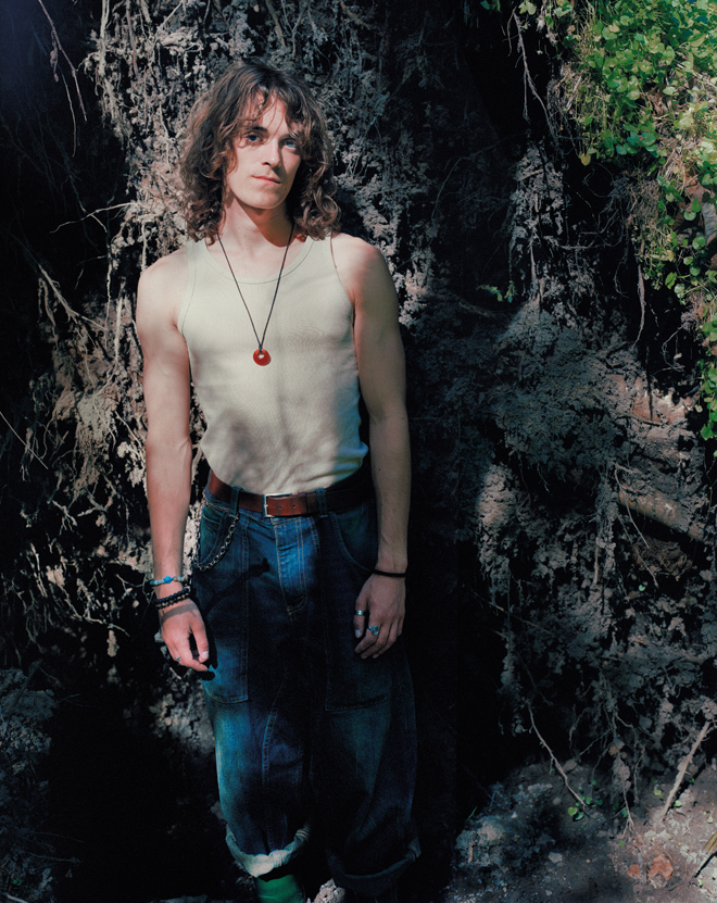 Young man with curly hair wearing a sleeveless beige shirt and baggy jeans stands by a tree root wall.