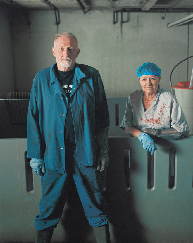 Two workers in blue uniforms and gloves standing in a dimly lit industrial room with concrete walls.