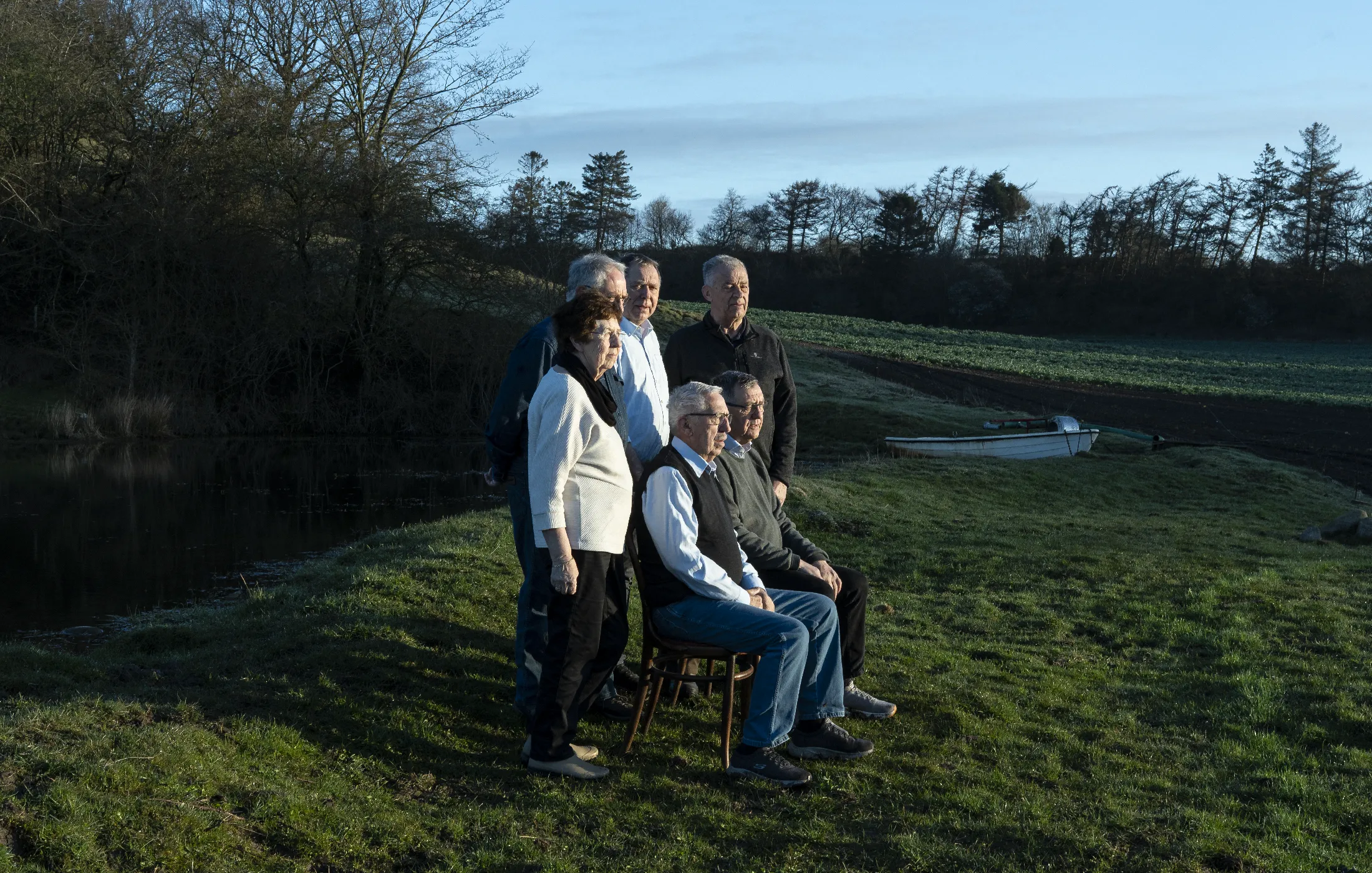 Group of six older adults outdoors by a lake with a small boat and trees in the background.