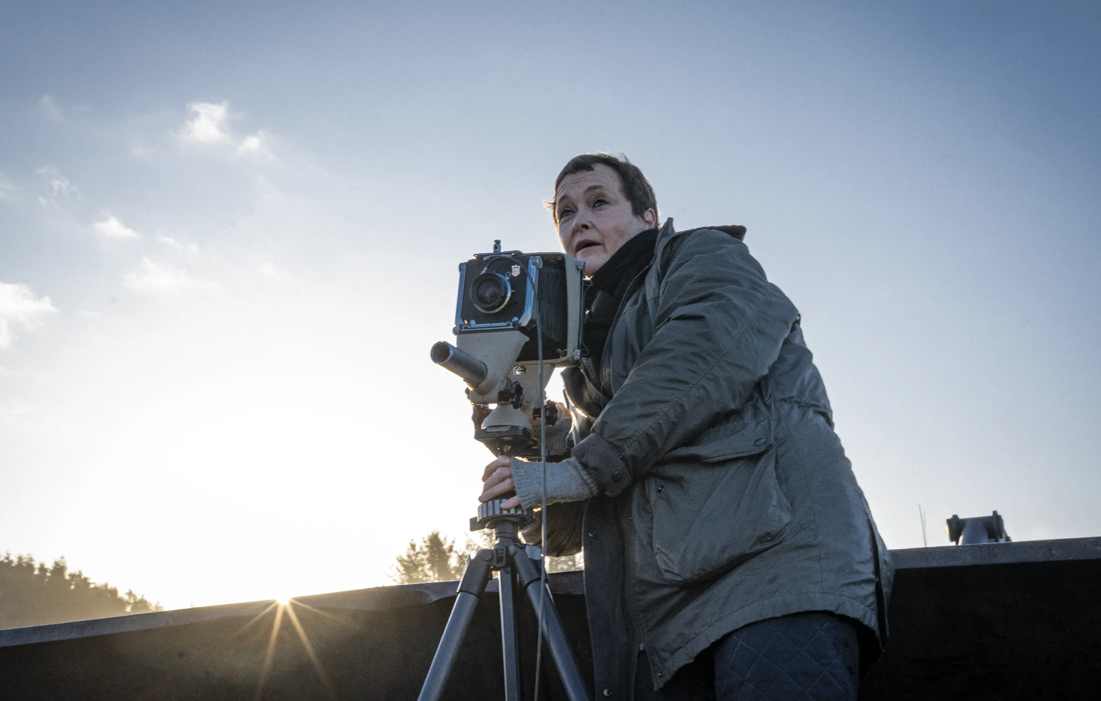 Person in a green jacket adjusting a large vintage camera on a tripod outdoors at sunset.
