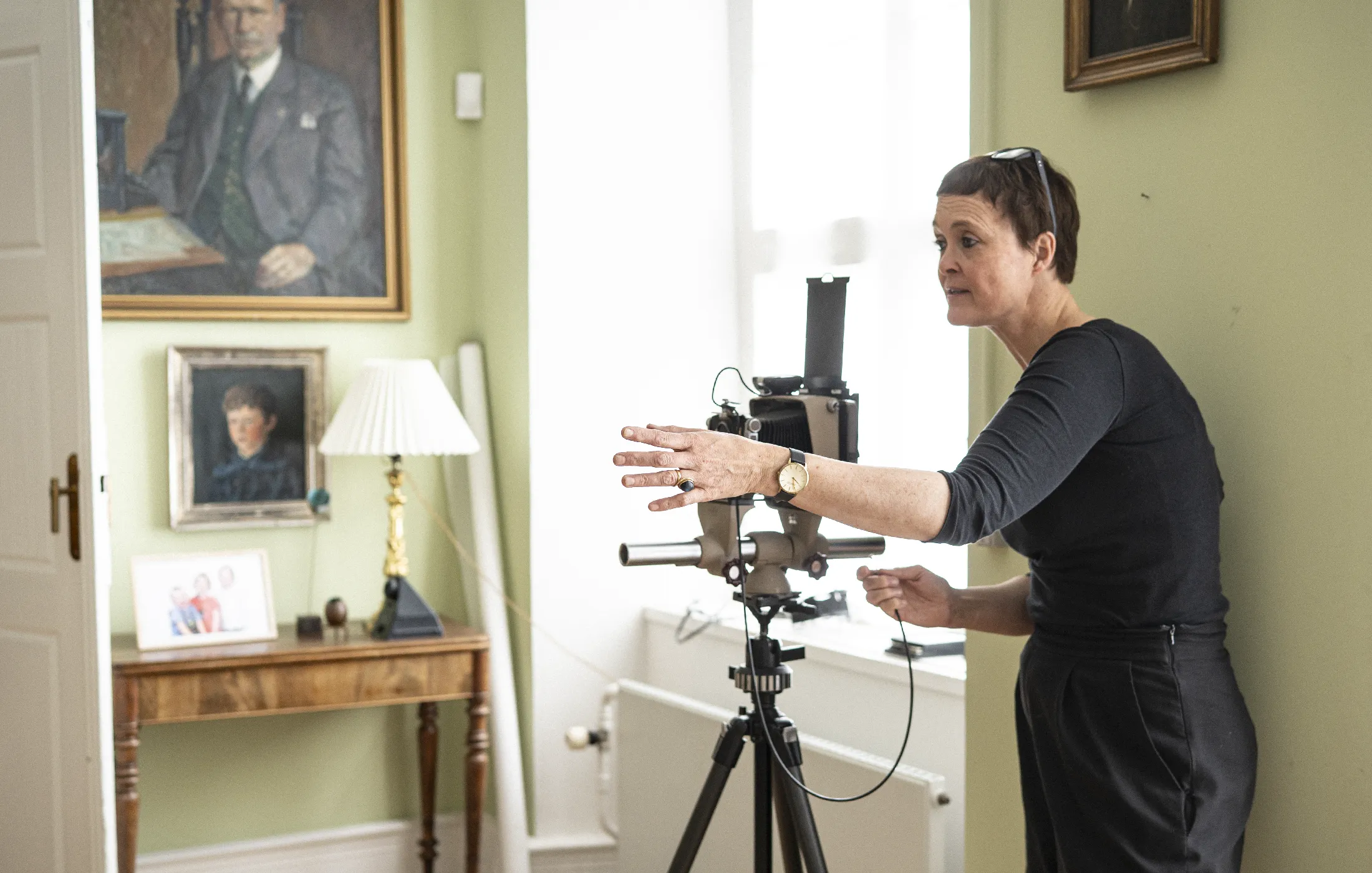 Person adjusting a large vintage camera on a tripod in a room with framed portraits and a lamp.