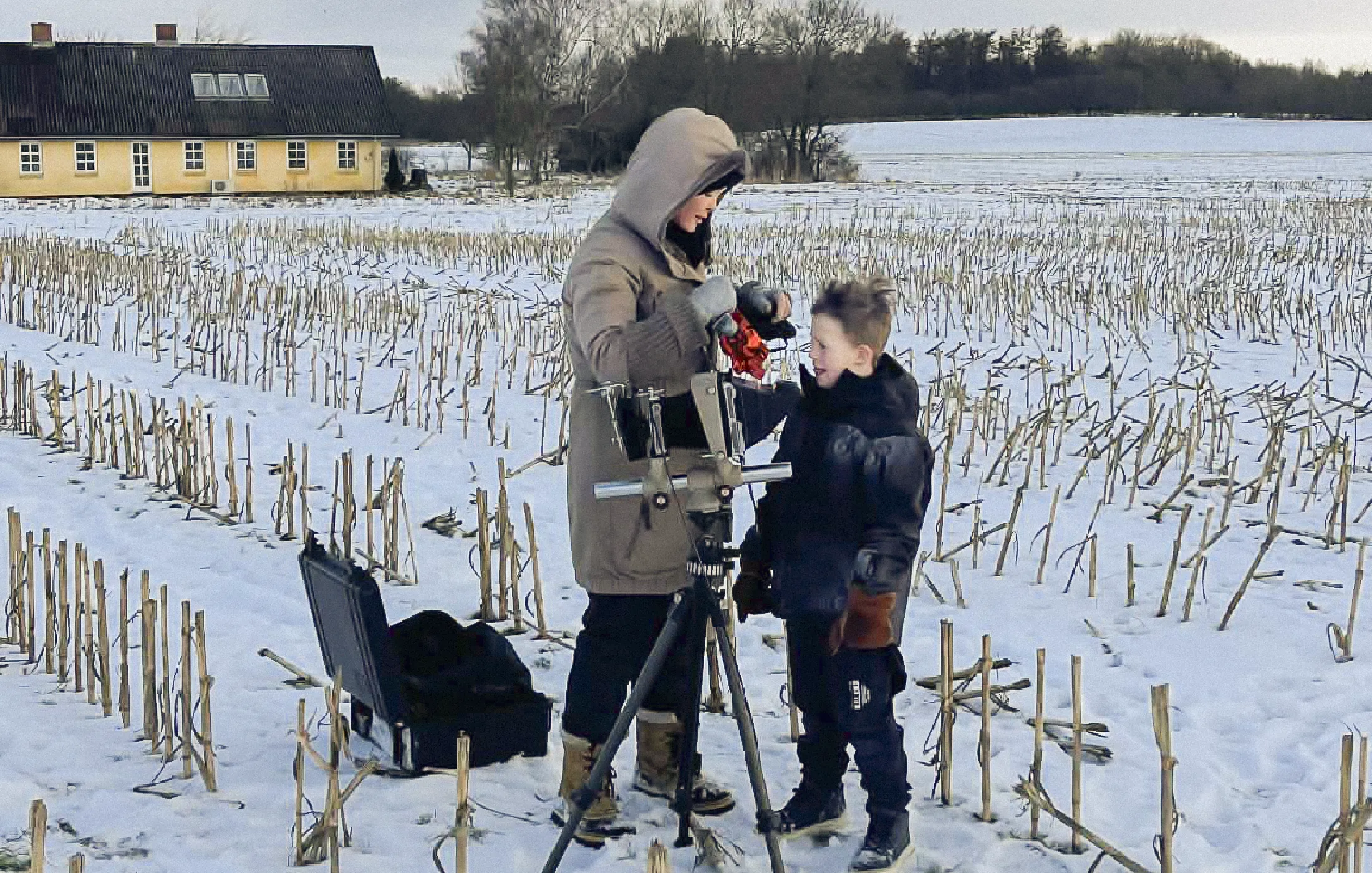 A woman and child in winter coats set up a telescope on tripod in a snowy field near a yellow house.