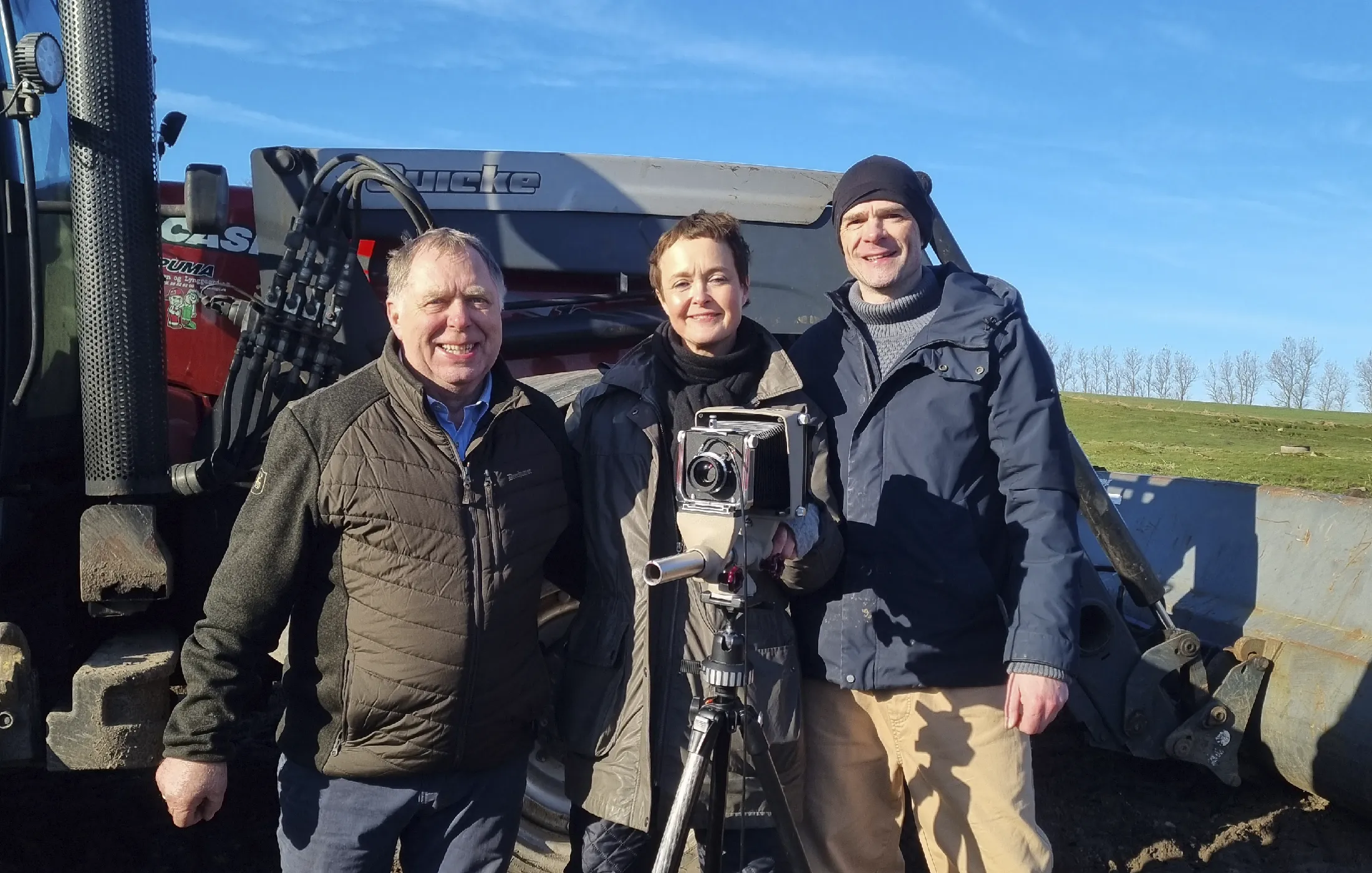 Three people standing outdoors in front of heavy machinery with a large camera on a tripod.