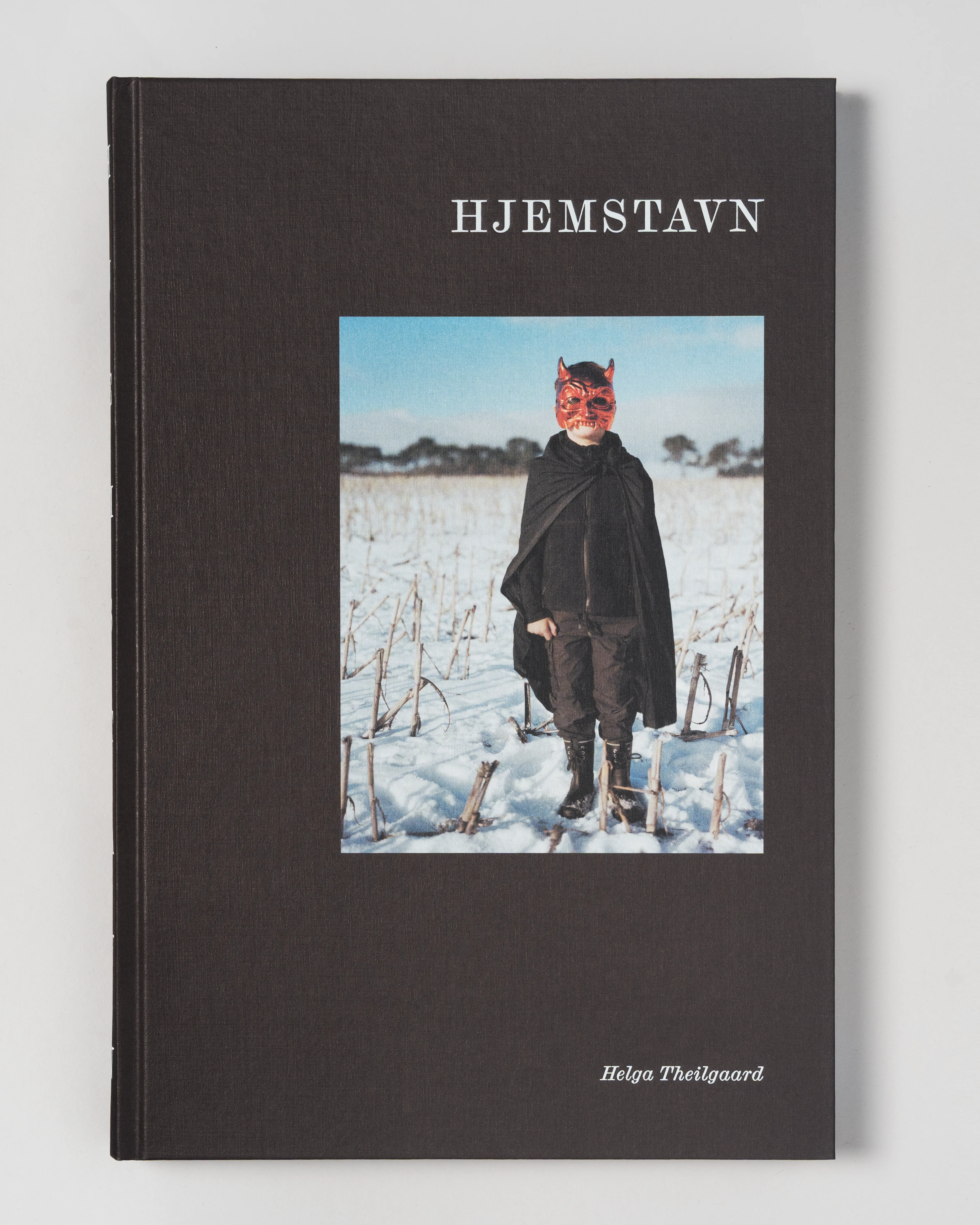 Child standing in snowy field wearing a red devil mask and black cape with boots.