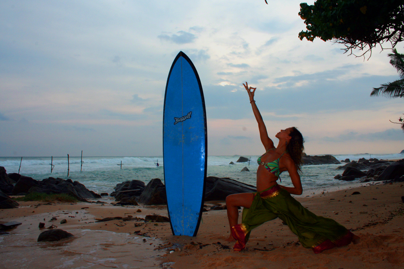 Mercedes in a warrior pose beside a blue surfboard on a Sri Lankan beach at dusk