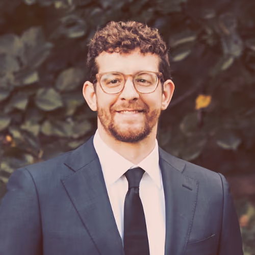 Smiling man with curly hair and glasses wearing a dark suit, white shirt, and black tie in front of foliage.
