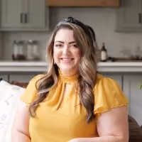 Smiling woman with wavy hair wearing a yellow blouse seated in a modern kitchen background.