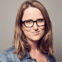 Woman with shoulder-length brown hair, black glasses, and denim jacket smiling against plain background.
