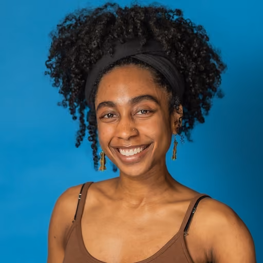 Smiling woman with curly hair, black headband, brown tank top, and dangling earrings on a blue background.