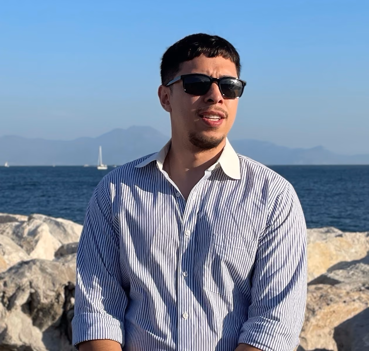 Man with sunglasses in a striped shirt standing by rocky shore with ocean and mountains behind him.