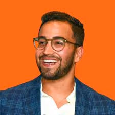 Smiling man with glasses and beard wearing a blue checkered blazer on an orange background.