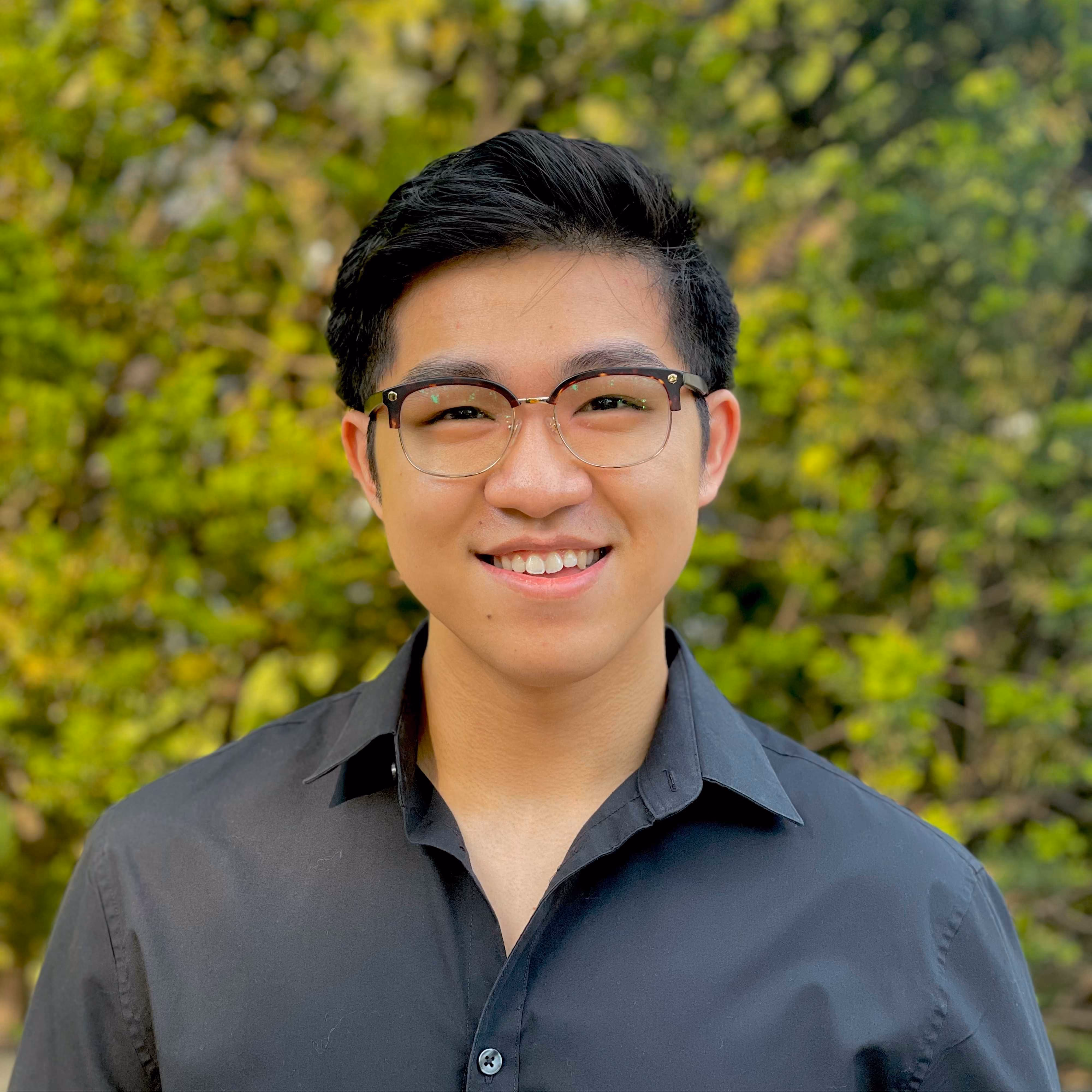 Smiling young man with glasses and black shirt standing outdoors with green foliage background.