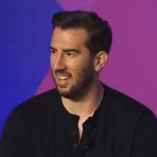 Man with dark hair and beard wearing a black shirt, smiling and talking against a purple background.