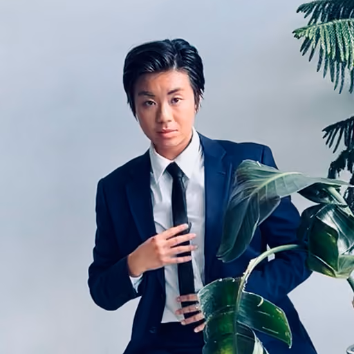Person wearing a dark suit and tie standing next to a large green plant against a plain background.