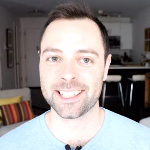 Smiling man with short dark hair and stubble wearing a light blue shirt indoors.