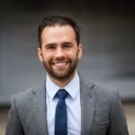 Smiling man with beard wearing a grey blazer, white shirt, and blue tie against a blurred background.