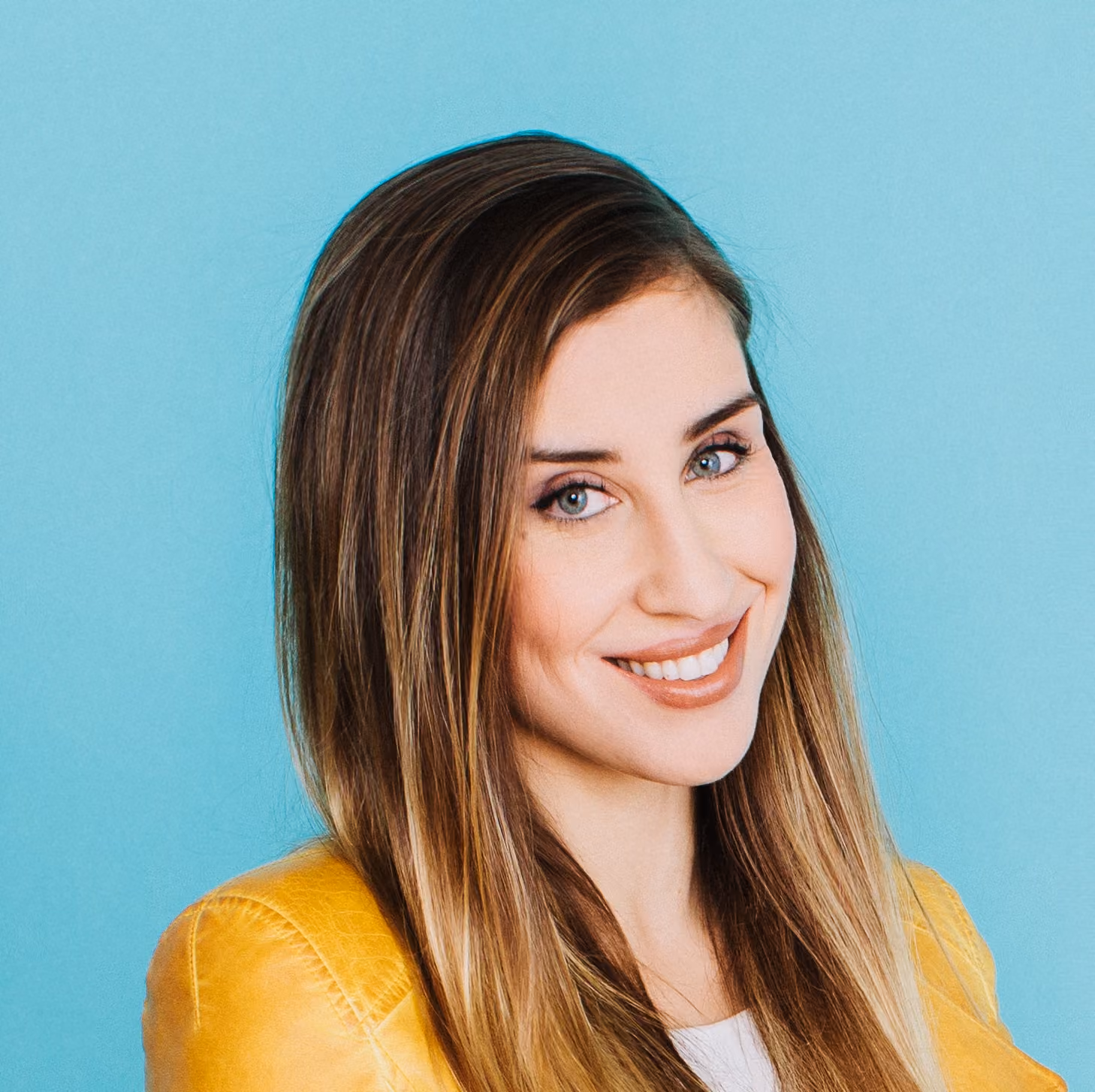 Smiling woman with long brown hair wearing a yellow jacket, against a blue background.