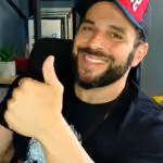 Bearded man smiling and giving a thumbs-up, wearing a black shirt and red cap indoors.
