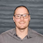 Smiling man with glasses and short hair wearing a gray shirt against a textured gray wall.