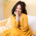 Smiling woman with curly hair wearing yellow dress sitting on sofa with patterned pillow.