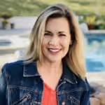 Smiling woman with blonde hair wearing a denim jacket and coral top outdoors by pool.