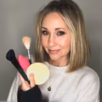 Woman with shoulder-length blonde hair holding various makeup brushes and a powder puff.
