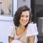 Smiling woman with wavy dark hair and tattoos wearing a white top sitting outdoors.