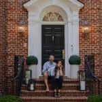 Couple sitting on brick steps outside a house with black door and white trim, smiling.