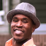 Smiling man wearing a gray fedora hat and a light-colored shirt outdoors.