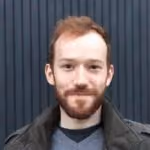 Young man with light brown hair and beard wearing a dark jacket and a gray sweater.