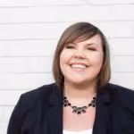 Smiling woman with short brown hair wearing a black blazer and necklace against a white brick wall.
