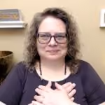 Person with curly hair and glasses holding both hands over their heart, smiling softly.