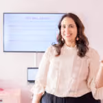 Smiling woman with dark hair and beige blouse standing in front of a blurred presentation screen.