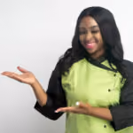 Smiling woman in green chef coat gesturing with both hands against a plain background.