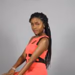 Woman with long braided hair posing confidently wearing a sleeveless coral dress.