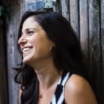 Smiling woman with long dark hair and hoop earrings standing by a wooden wall.