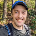 Smiling man wearing cap and backpack in a wooded outdoor setting with sunlight filtering through trees.