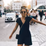 Smiling woman in black dress with sunglasses walking on a city street with cars and people.