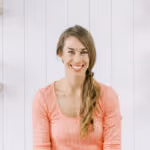 Smiling woman with long side braid wearing a coral top against a white wooden wall.