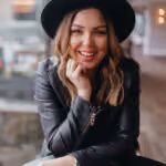 Smiling woman with blonde hair wearing a black hat and leather jacket, resting her chin on her hand.