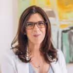 Woman with brown hair wearing glasses and a white blazer smiling indoors with artwork in background.