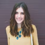 Smiling woman with shoulder-length brown hair wearing a mustard sweater and teal necklace against a brown wall.
