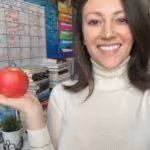 Woman in white turtleneck smiling and holding a red apple in one hand in a room with colorful shelves.