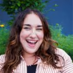 Smiling woman with curly hair wearing a striped shirt in front of green plants and a blue wall.