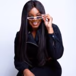 Woman with long straight hair smiling, wearing sunglasses and a black outfit against a white background.