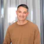 Smiling man with short dark hair wearing a brown sweater standing indoors with curtains in the background.