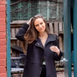 Woman with long brown hair in black coat standing outdoors by a green door and red wall.