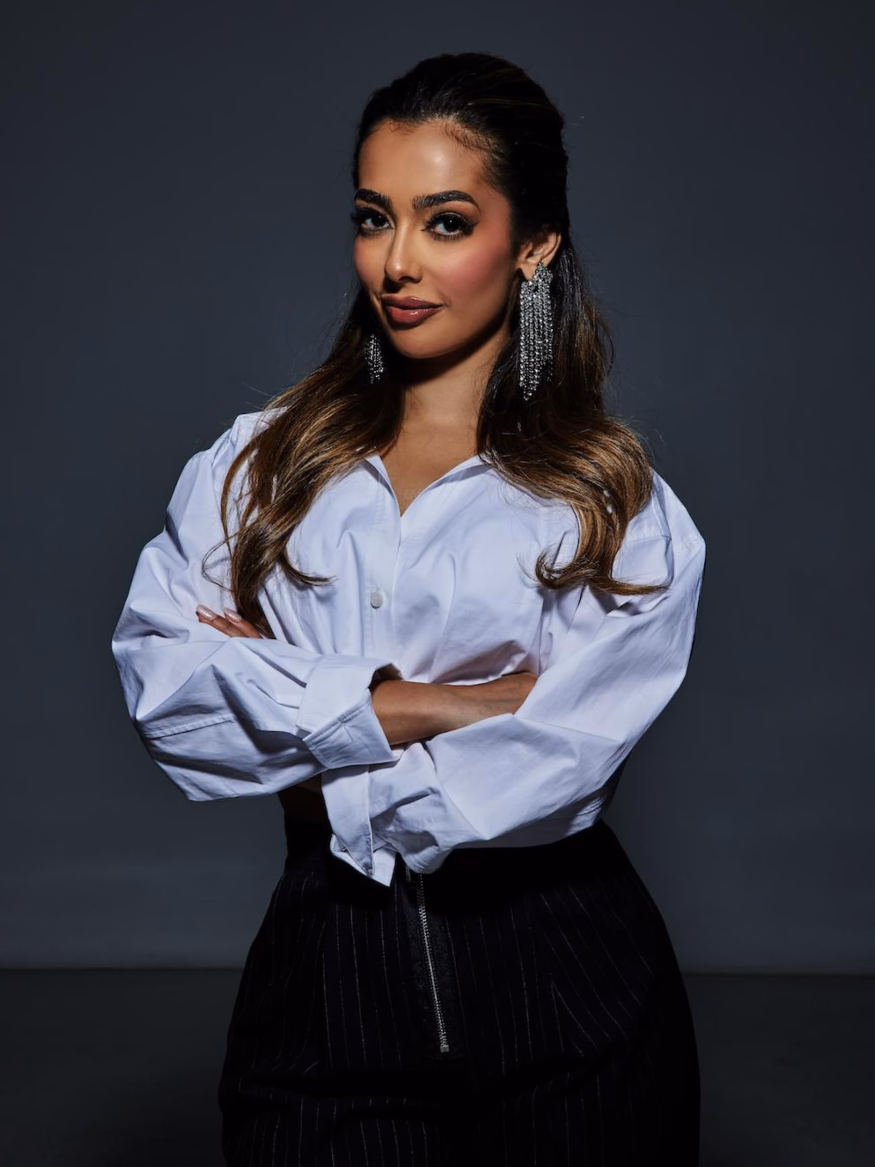 Confident woman with long hair, white blouse, and statement earrings stands with arms crossed.