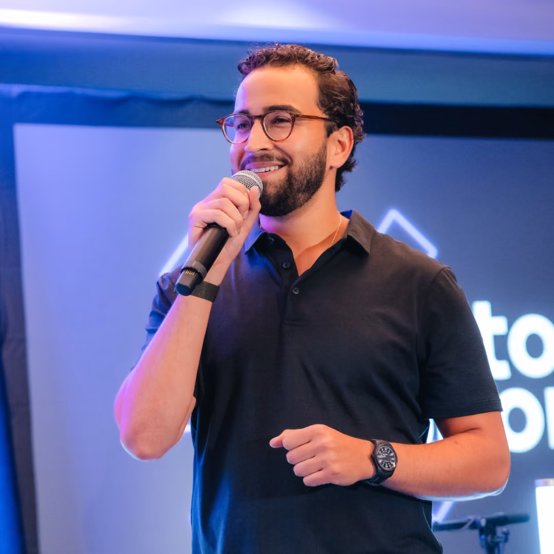 Smiling man with glasses and beard speaking into a microphone on stage with blue background.